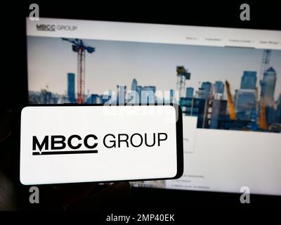 Personne tenant le téléphone cellulaire avec le logo de la société de produits chimiques de construction MBCC Group sur l'écran en face de la page Web d'affaires. Mise au point sur l'affichage du téléphone. Banque D'Images