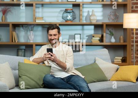 Jeune homme souriant dans une chemise beige assis sur un canapé à la maison pendant la quarantaine. Tenant le téléphone entre ses mains, il écrit des messages avec sa petite amie, son amant, ses amis, ses parents, sa famille. Banque D'Images