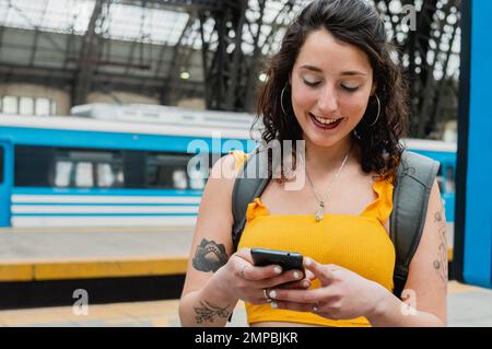 jeune femme latine d'origine argentine, debout sur la plate-forme du terminal ferroviaire, se réjouit de lire les notifications sur son téléphone et de vérifier Banque D'Images