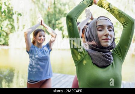 Groupe d'adolescents est engagé dans la forme physique sur la jetée au bord du lac ou de la rivière. Les jeunes filles et les filles positives vont dans le sport. Musulman dans un hijab, afro-américain A. Banque D'Images