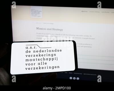 Personne tenant un téléphone portable avec le logo de la compagnie d'assurance néerlandaise ASR Nederland NV à l'écran en face de la page Web des affaires. Mise au point sur l'affichage du téléphone. Banque D'Images