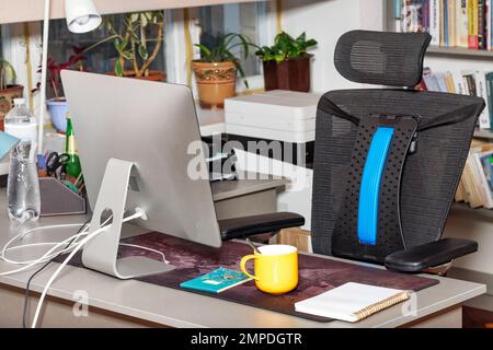 Un lieu de travail avec un moniteur, une chaise confortable, un bloc-notes et une tasse jaune sur un fond flou près de la fenêtre. Banque D'Images