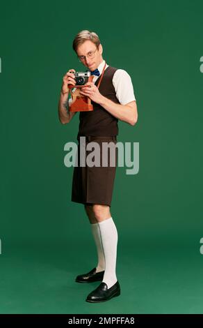 Prendre des photos avec un appareil photo vintage. Portrait de l'homme dans des vêtements classiques élégants, short et gilet posé sur fond vert studio. Concept de Banque D'Images