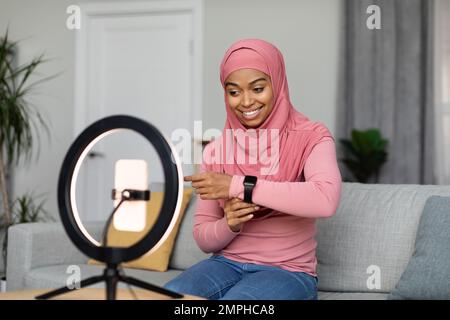 Femme noire excitée dans le hijab faisant une vue d'ensemble des appareils et montrant la montre intelligente, l'enregistrement vidéo sur le téléphone portable à la maison Banque D'Images