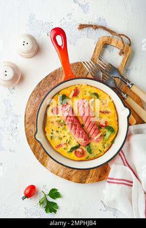 Omelette ou frittata avec deux saucisses, fromage fondu, tomates cerises, brocoli et persil sur poêle rouge ou poêle en fer sur fond blanc en béton Banque D'Images
