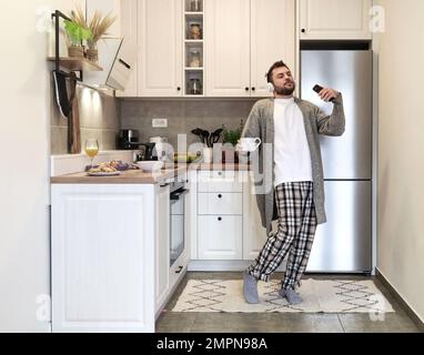 Homme barbu adulte avec un casque tenant une tasse à café et un téléphone portable dans ses mains dansant et appréciant la musique avec les yeux fermés dans la cuisine domestique dans Banque D'Images