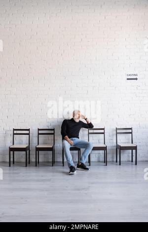 homme aux cheveux gris lassés assis sur une chaise près du mur blanc et attendant la coulée Banque D'Images