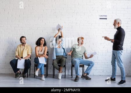 le directeur artistique barbu montre la bonne actrice afro-américaine qui tient le scénario à mains levées tout en étant assise près de chercheurs d'emploi interraciaux Banque D'Images