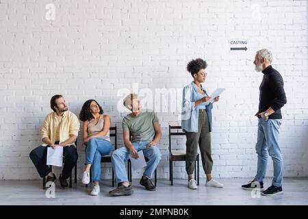 un directeur artistique barbu debout avec les mains dans les poches près d'une femme afro-américaine lisant un scénario et des acteurs multiethniques Banque D'Images