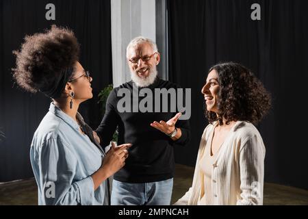 positif gris cheveux producteur pointant avec la main tout en parlant à gaies actrices interraciales dans le théâtre Banque D'Images