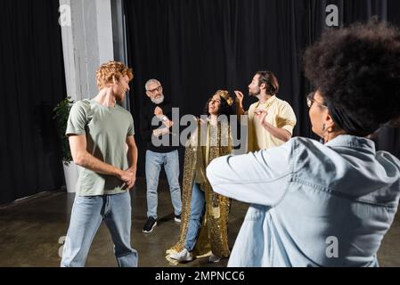 joyeuse femme multiraciale en costume de reine souriant près des acteurs interraciaux et du directeur artistique à cheveux gris. Traduction de tatouage: om, shanti, paix Banque D'Images