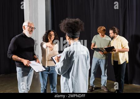 femme multiraciale souriante pointant de la main près du directeur artistique barbu et de l'actrice afro-américaine sur la scène théâtrale Banque D'Images