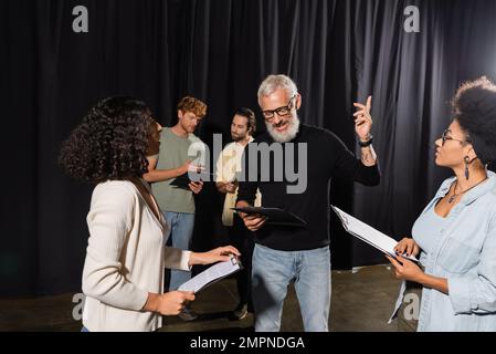 le directeur artistique barbu pointe avec le doigt tout en lisant un scénario près d'actrices multiethniques pendant la leçon de compétences d'action Banque D'Images