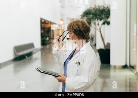 Vue latérale d'une femme médecin portant des lunettes de vue tout en tenant le dossier à l'hôpital Banque D'Images