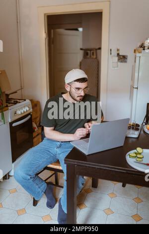 Homme utilisant un ordinateur portable sur la table à manger assis dans la cuisine à la maison Banque D'Images
