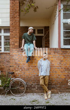 Homme regardant un petit ami penché sur le mur de briques à vélo à l'extérieur de la maison Banque D'Images