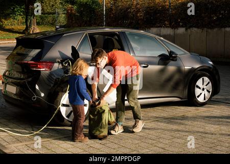 Père cherchant dans un sac à dos avec fille debout près de la voiture électrique en charge à la gare Banque D'Images