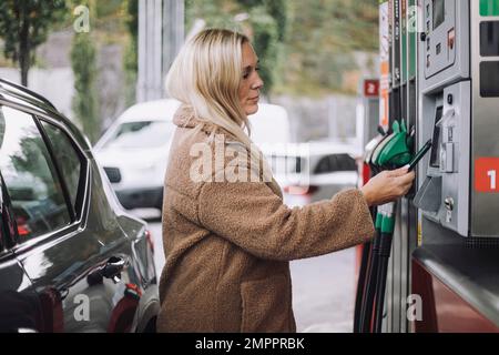 Vue latérale d'une femme blonde qui effectue le paiement via un smartphone en se tenant à la station-service Banque D'Images