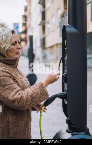 Femme mature qui paie par portefeuille numérique à la station de charge de véhicule électrique Banque D'Images