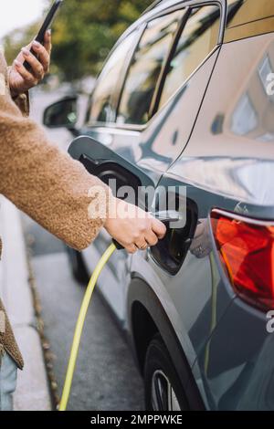 Main de la femme qui branche le chargeur dans la voiture électrique à la station Banque D'Images