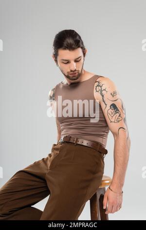 Un homme stylé tatoué dans un débardeur marron, assis sur une chaise isolée sur un gris Banque D'Images