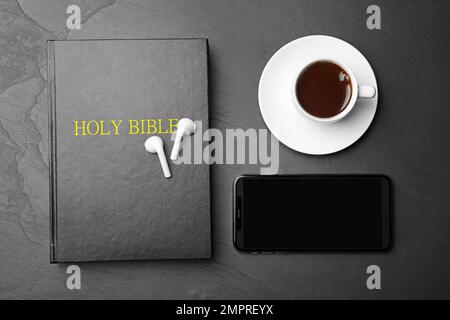 Bible, téléphone, tasse de café et écouteurs sur fond noir, plat. Livre audio religieux Banque D'Images
