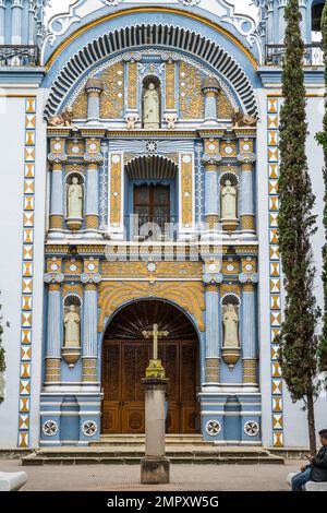 Croix en pierre devant la façade peinte et colorée de l'église Saint-Domingue de Guzman à Ocotlan de Morelos, Oaxaca, Mexique. Banque D'Images