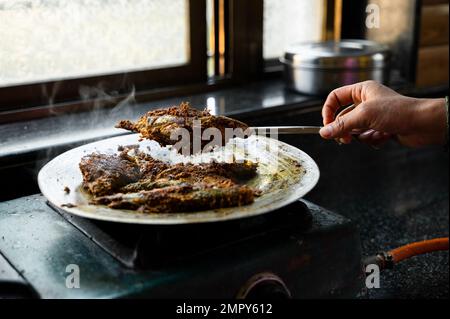 Maquereau indien poisson mariné avec des épices friture sur métal sur l'ustensile en aluminium et un morceau de poisson soulevé à l'aide d'une cuillère avec la main de femme. Banque D'Images