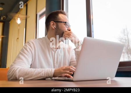 Homme d'affaires élégant et réfléchi d'apparence européenne dans des lunettes, avec une barbe, dans une chemise blanche qui donne sur la fenêtre distrait du travail sur les genoux Banque D'Images