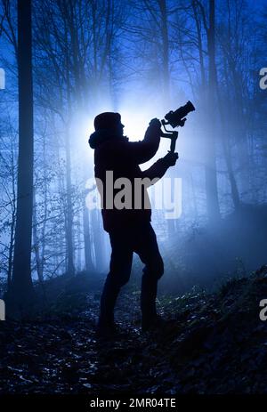 Silhouette d'un vidéaste dans une forêt brumeuse, rétro-éclairé de façon spectaculaire avec une couleur bleu froid et des rayons de lumière derrière lui Banque D'Images