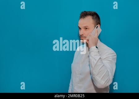 Élégant homme d'affaires sérieux homme d'affaires d'apparence européenne dans une chemise blanche parle sur un smartphone contre le fond bleu du mur. Banque D'Images