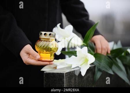 Femme avec des bougies et des nénuphars blancs près de la pierre tombale de granit noir à l'extérieur, gros plan. Cérémonie funéraire Banque D'Images