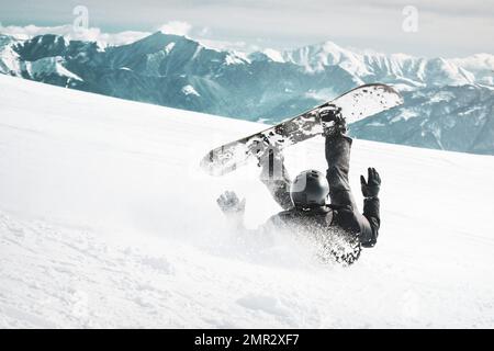 Le snowboarder tombe sur le dos avec des projections de neige sur la piste de ski de fond enneigée et le télésiège ancien en arrière-plan. Montagnes du Caucase en hiver ensoleillé Banque D'Images