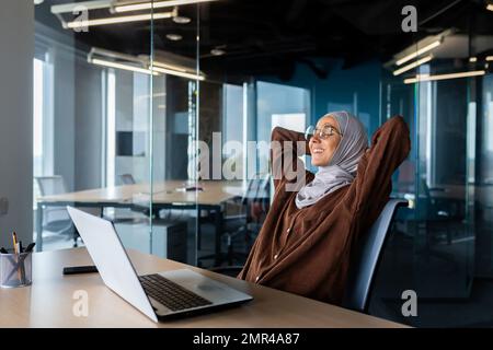 Femme d'affaires dans le hijab se reposant, femme arabe dans le hijab avec ses mains derrière sa tête et souriant avec les yeux fermés, femme en lunettes rêvant de résultats futurs, de bonnes réalisations et de succès. Banque D'Images