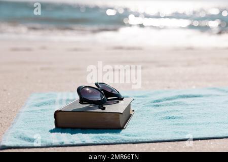 Gros plan du livre avec lunettes de soleil et serviette bleue sur la plage de sable face à la mer pendant la journée ensoleillée Banque D'Images