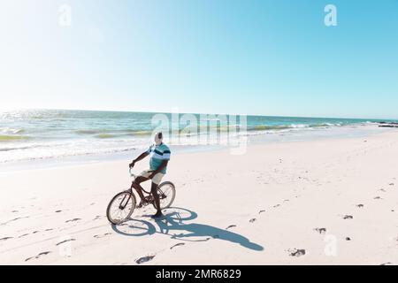 Homme afro-américain senior à vélo sur une plage de sable contre la mer et ciel clair par beau temps Banque D'Images