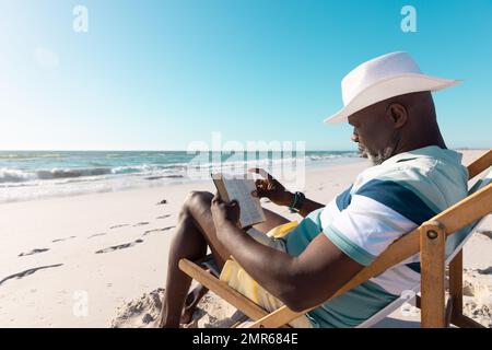 Vue latérale d'un homme senior afro-américain portant un chapeau de lecture sur un transat à la plage sous le ciel Banque D'Images