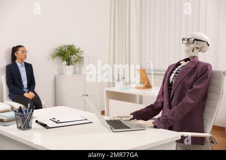 Squelette humain en costume utilisant un ordinateur portable à la table et une femme attendant sur une chaise dans le bureau Banque D'Images