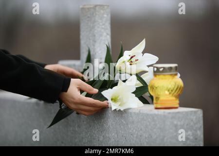 Femme tenant des lys blancs près de la tombe en granit gris clair avec bougie à l'extérieur, gros plan. Cérémonie funéraire Banque D'Images