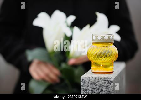 Femme tenant des lys blancs près de la tombe en granit gris à l'extérieur, se concentrer sur la bougie. Cérémonie funéraire Banque D'Images