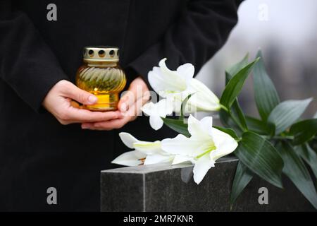 Femme avec des bougies et des nénuphars blancs près de la pierre tombale de granit noir à l'extérieur, gros plan. Cérémonie funéraire Banque D'Images