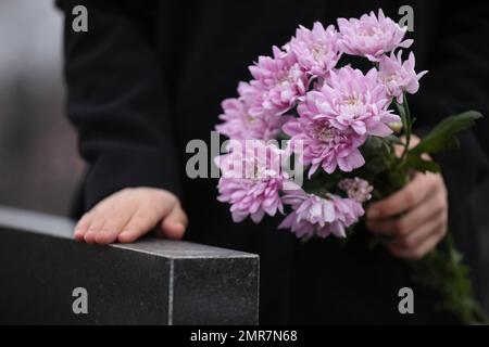 Femme tenant des fleurs de chrysanthème près de la pierre tombale de granit noir à l'extérieur, gros plan. Cérémonie funéraire Banque D'Images