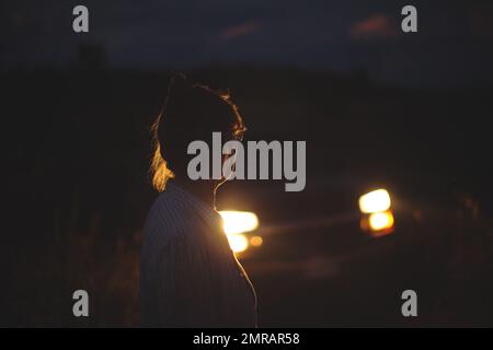 Une femme debout sur un terrain contre une voiture avec les phares allumés Banque D'Images