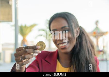 Une femme négociante afro-américaine attirante sourit en tenant une pièce de monnaie en bitcoin. Point de mire sur la pièce Banque D'Images
