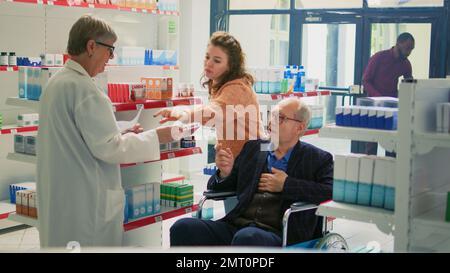 Pharmacien donnant des pilules du papier d'ordonnance à l'homme avec un handicap, personne en fauteuil roulant venant à la pharmacie avec un gardien. Aide sociale aidant les adultes âgés avec des traitements et des vitamines. Banque D'Images