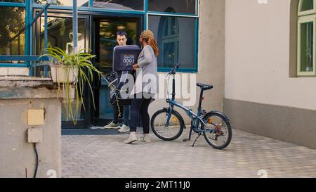 L'employé du service de livraison express livre le déjeuner dans le sac à dos, donnant la commande de fastfood à la porte d'entrée. Client souriant prenant un repas à emporter au restaurant à l'extérieur, coursier avec forfait sur vélo. Banque D'Images