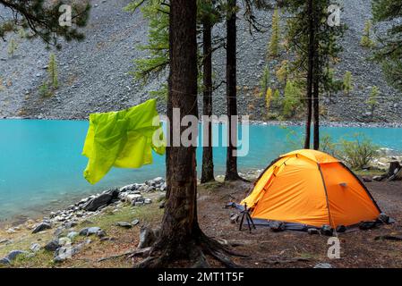 Une tente orange se dresse dans des épinettes sur la rive du lac turquoise de haute altitude Shavlinskoye en Altai pendant la journée avec un imperméable qui s'assèche Banque D'Images