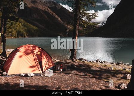 Une tente orange se dresse dans les arbres sur la rive du lac turquoise de haute altitude Shavlinskoye en Altai dans l'après-midi après une pluie. Banque D'Images