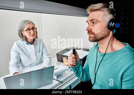 Un patient de sexe masculin porte un casque d'audiologie appuyant sur le bouton de réponse pendant les tests audiométriques dans une cabine d'audiologie insonorisée. Tests auditifs Banque D'Images