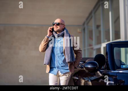 Photo d'un homme d'âge moyen portant des vêtements décontractés et utilisant un téléphone portable tout en se tenant debout à côté de sa voiture. Banque D'Images